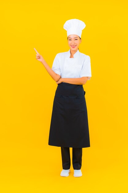 Ritratto bella giovane donna asiatica in chef o cuoco uniforme con cappello su sfondo giallo isolato