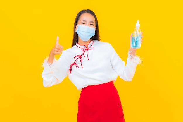 Ritratto bella giovane donna asiatica indossa maschera e usa gel alcolico per proteggere covid19 su giallo