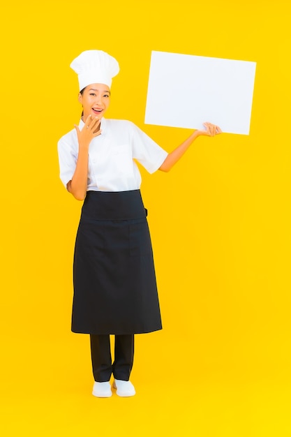 Ritratto bella giovane donna chef asiatica con cartellone vuoto bianco su sfondo giallo isolato