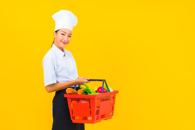 Ritratto bella giovane donna chef asiatica con cesto della spesa dal supermercato su sfondo giallo isolato