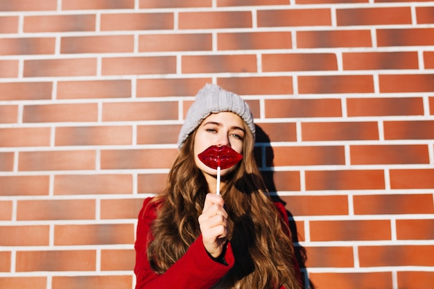 Ritratto bella ragazza bruna con labbra lecca-lecca sul muro esterno. Indossa cappello lavorato a maglia, cappotto rosso.