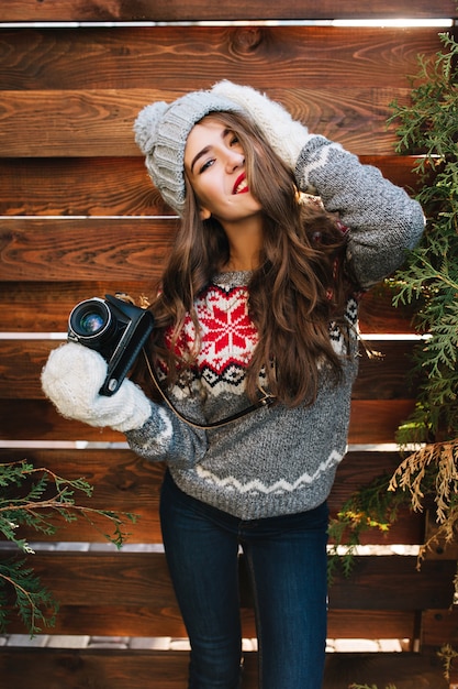 Ritratto bella ragazza con capelli lunghi in cappello lavorato a maglia e guanti che tengono la macchina fotografica su legno. Lei sta sorridendo .