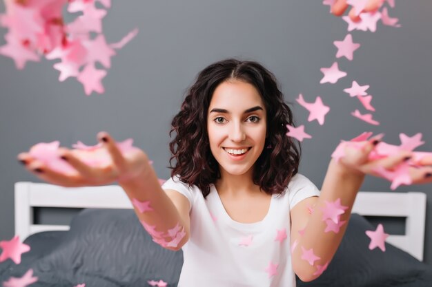 Ritratto carino allegro abbastanza giovane donna in pigiama con i capelli ricci bruna divertendosi sul letto. Allungando le mani con orpelli rosa, espressione di felicità