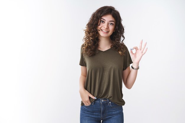 Ritratto carino armeno dai capelli ricci donna in t-shirt verde oliva mostra ok gesto pensiero vestito non male d'accordo dire ok, sorridente dare approvazione confermare che tutto va come previsto, in piedi sfondo bianco