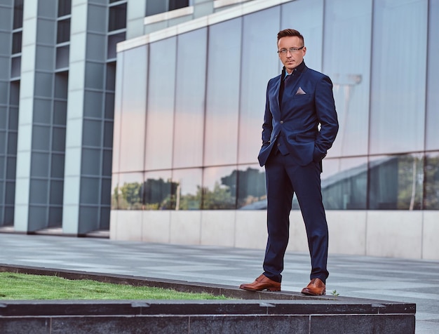 Ritratto completo del corpo di un uomo sicuro di sé in abito elegante in piedi con le mani in tasca su uno sfondo di grattacielo.