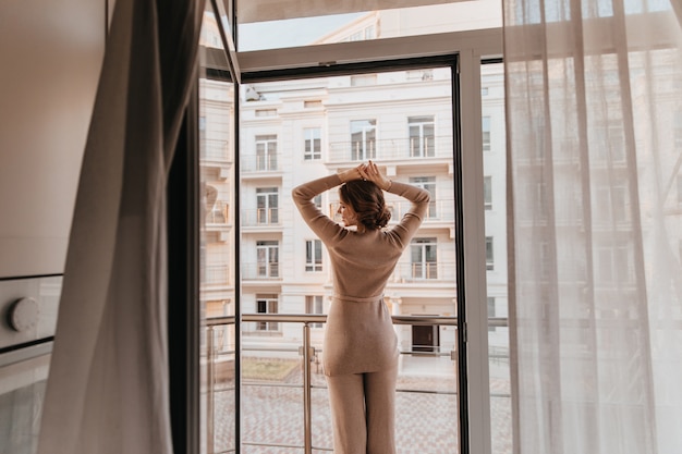 Ritratto dal retro della donna rilassata in abito marrone. Foto di signora alla moda felice che posa vicino alla grande finestra.
