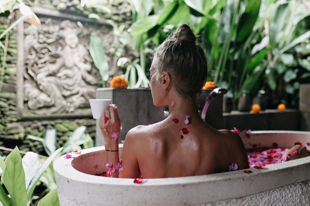 Ritratto dal retro della graziosa donna europea che cattura bagno con petali di rosa e degustazione di tè.