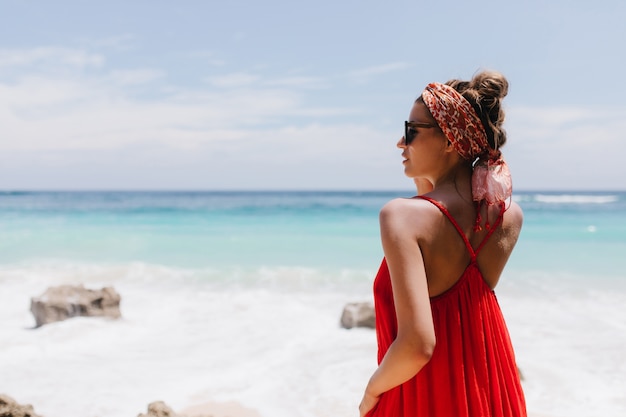 Ritratto dal retro della ragazza spensierata con la pelle abbronzata guardando l'orizzonte. Foto del modello femminile caucasico felice in abbigliamento rosso agghiacciante sulla costa dell'oceano e godendo della vista.