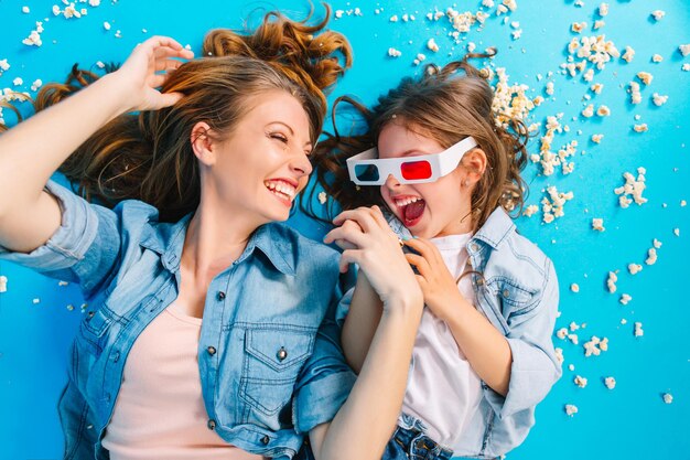 Ritratto dall'alto tempo felice insieme di bella madre che si diverte con la figlia sul pavimento blu. Sdraiarsi nei popcorn, godersi la felicità familiare, esprimere vere emozioni positive