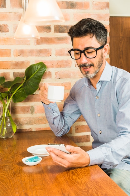 Ritratto degli occhiali d&#39;uso di un uomo che tengono la tazza di caffè che esamina smartphone