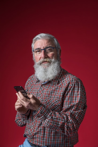 Ritratto degli occhiali d&#39;uso di un uomo senior che tengono Smart Phone a disposizione contro il contesto rosso