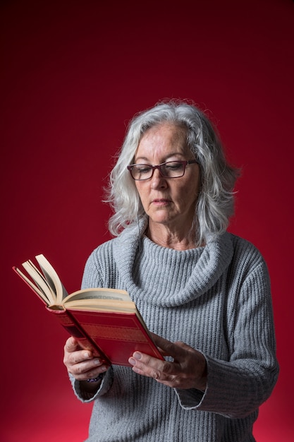 Ritratto degli occhiali d&#39;uso di una donna senior che leggono il libro che sta contro il fondo rosso