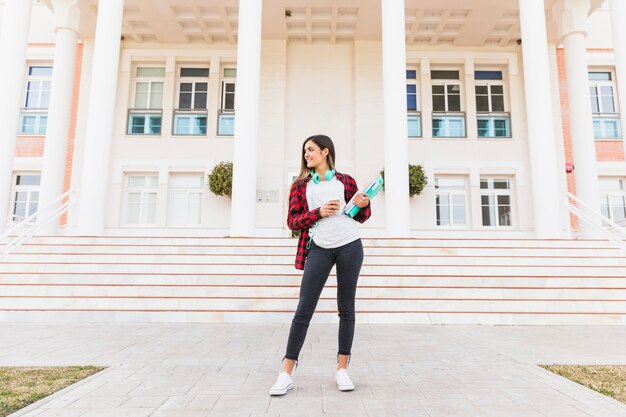 Ritratto dei libri sorridenti della tenuta della studentessa e della tazza di caffè asportabile che stanno davanti all&#39;edificio universitario