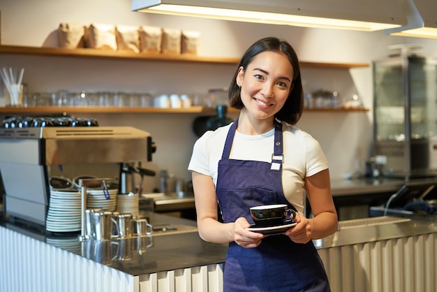 Ritratto del barista femminile sorridente asiatico che indossa il grembiule del caffè e che tiene la tazza di caffè che prende il cliente