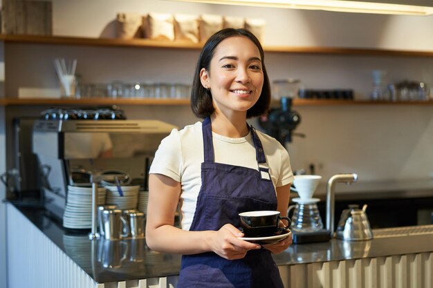 Ritratto del barista femminile sorridente asiatico che indossa il grembiule del caffè e che tiene la tazza di caffè che prende il cliente