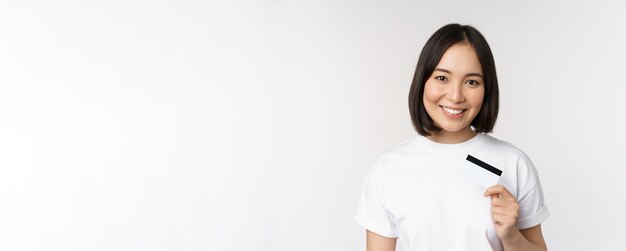 Ritratto del cliente coreano sorridente della banca della ragazza che mostra la carta di credito con la faccia felice che si leva in piedi contro il whi