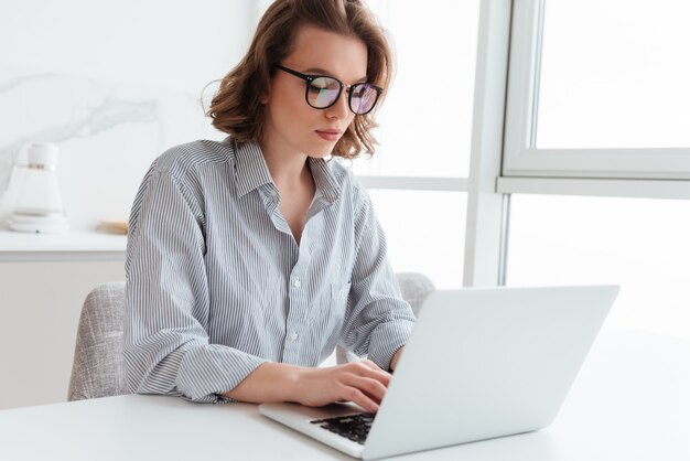 Ritratto del email mandante un sms della giovane donna elegante sul computer portatile mentre sedendosi alla tavola nella stanza leggera