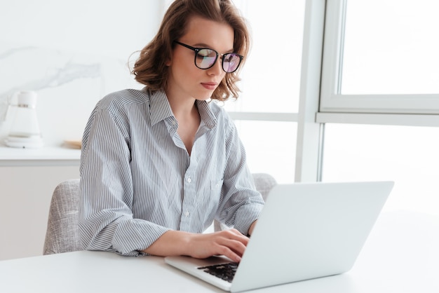 Ritratto del email mandante un sms della giovane donna elegante sul computer portatile mentre sedendosi alla tavola nella stanza leggera