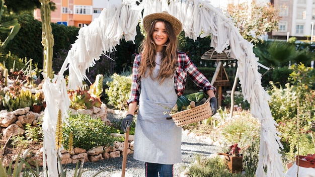 Ritratto del giardiniere femminile sorridente che sta con gli strumenti di giardinaggio e canestro nel giardino