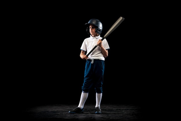 Ritratto del giocatore di baseball del bambino del ragazzo nella pratica uniforme della formazione isolata sopra il fondo nero dello studio