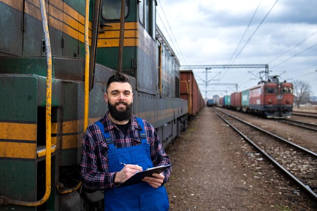 Ritratto del macchinista del treno in piedi dalla locomotiva alla stazione ferroviaria e tenendo il programma di partenza