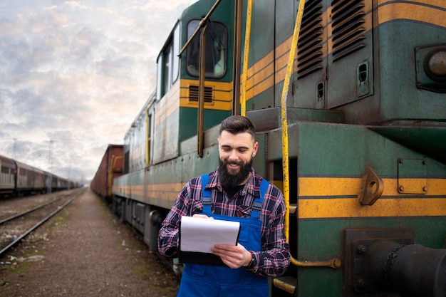 Ritratto del macchinista del treno in piedi dalla locomotiva alla stazione ferroviaria e tenendo il programma di partenza