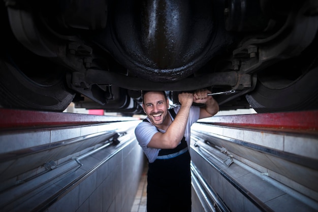 Ritratto del meccanico sorridente del veicolo che tiene la chiave e lavora sotto il camion nell'officina di riparazione del veicolo