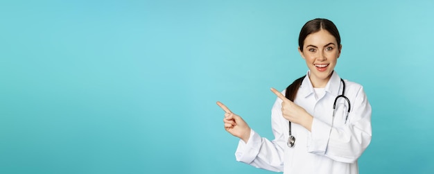 Ritratto del medico sorridente della ragazza dell'operaio medico in camice bianco con lo stetoscopio che punta le dita a sinistra