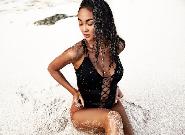 ritratto del modello di bella donna caucasica preso il sole con i capelli lunghi scuri in costume da bagno nero in posa sulla spiaggia estiva con sabbia bianca