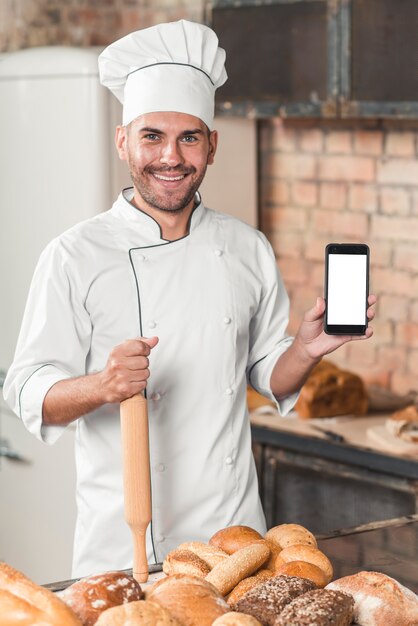 Ritratto del panettiere maschio che mostra cellulare in bianco con i pani al forno