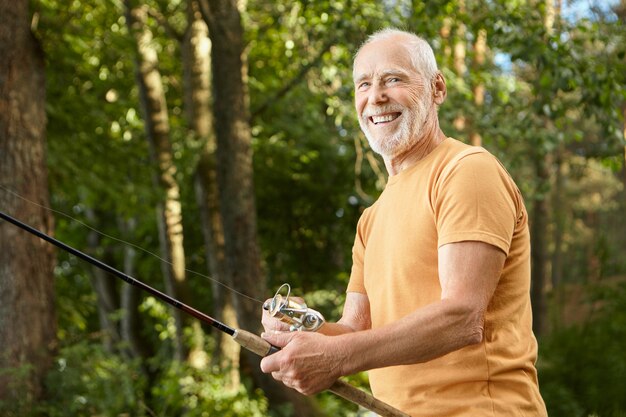 Ritratto del pensionato maschio caucasico barbuto sorridente sano in maglietta che propone all'aperto con gli alberi verdi che tengono la canna da pesca, godendo di pesca. Ricreazione, tempo libero e concetto di natura
