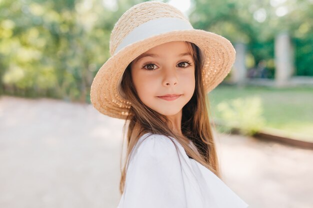 Ritratto del primo piano del bambino meraviglioso con gli occhi marroni lucidi che guardano con interesse. Bambina entusiasta in cappello di paglia vintage decorato con nastro in posa durante il gioco nel parco.