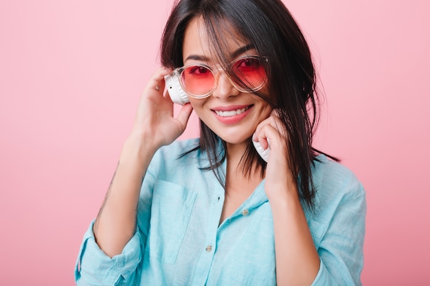 Ritratto del primo piano del modello femminile ispanico elegante indossa occhiali da sole colorati e camicia di cotone. Ispirata donna latina in abiti blu che si gode una buona canzone.