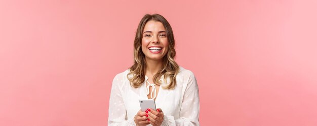 Ritratto del primo piano dell'affascinante ragazza bionda adorabile che utilizza il telefono cellulare che invia un sms all'ordine dell'amico nel negozio online utilizzando l'applicazione per smartphone ridendo e sorridendo soddisfatto dello sfondo rosa
