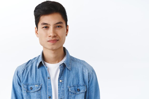 Ritratto del primo piano dell'uomo asiatico alla moda bello dei pantaloni a vita bassa con la macchina fotografica compiaciuta sorridente dell'orecchino che sembra studente entusiasta e in buona salute che cerca lavoro dopo il fondo bianco di graduazione