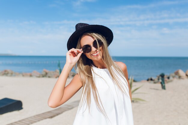 Ritratto del primo piano della bella ragazza bionda con i capelli lunghi è in piedi sulla spiaggia vicino al mare. Sta sorridendo alla telecamera.