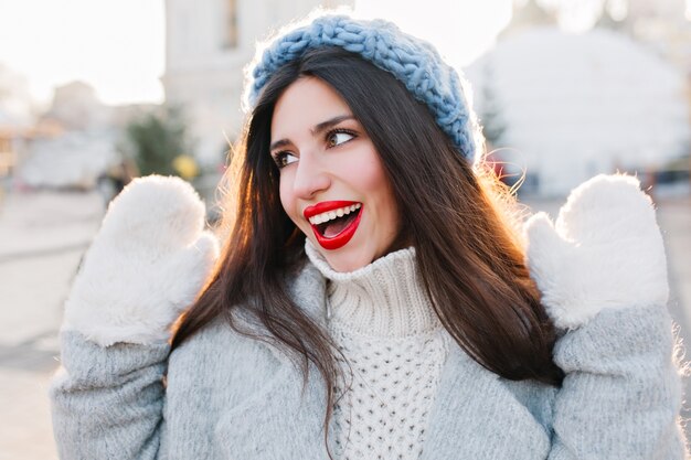 Ritratto del primo piano della bella ragazza con i capelli lunghi neri in posa con le mani in una giornata fredda. Foto all'aperto di carina signora europea con cappello blu e guanti bianchi che si godono il fine settimana invernale.