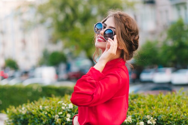 Ritratto del primo piano della bella ragazza in occhiali da sole in posa per la fotocamera nel parco. Indossa una camicetta rossa e una bella acconciatura. Sta guardando lontano.