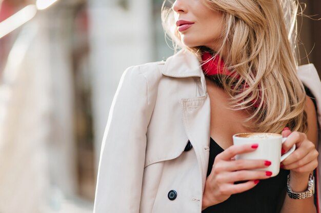 Ritratto del primo piano della donna bionda raffinata in cappotto che tiene tazza bianca con bevanda. Affascinante signora bionda che beve caffè in una giornata fredda e distoglie lo sguardo.