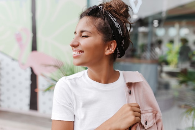 Ritratto del primo piano della donna dai capelli scuri abbronzata con panino alla moda, sorridente contro il muro del bar con fenicotteri dipinti