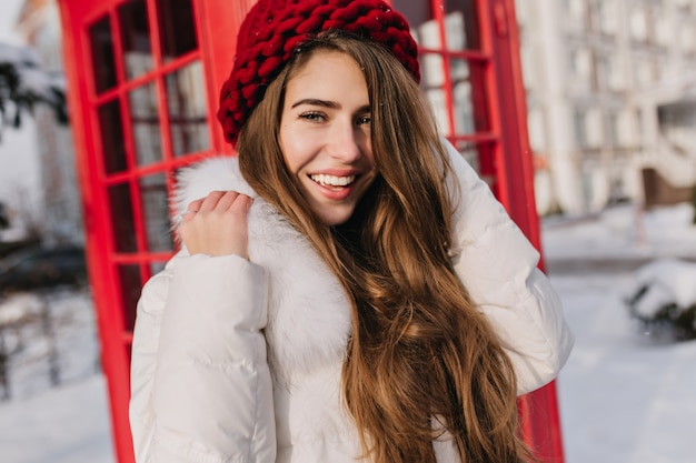 Ritratto del primo piano della donna felice con capelli castani lucidi che posano accanto alla cabina telefonica rossa. Foto all'aperto di una splendida modella in berretto lavorato a maglia che si gode la gelida mattina in Inghilterra.