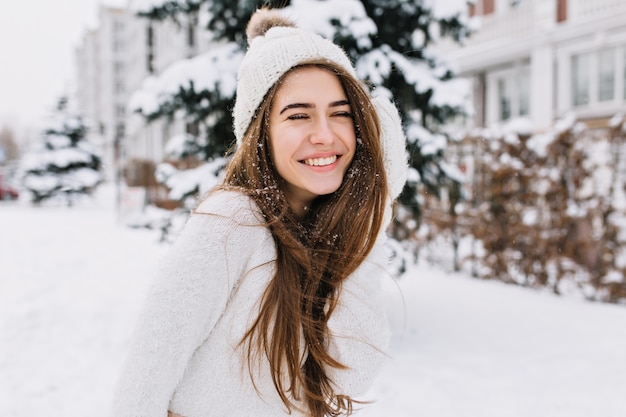 Ritratto del primo piano della donna felice in maglione di lana che gode dei momenti di inverno. Foto all'aperto della signora che ride dai capelli lunghi in cappello lavorato a maglia divertendosi nella mattina nevosa