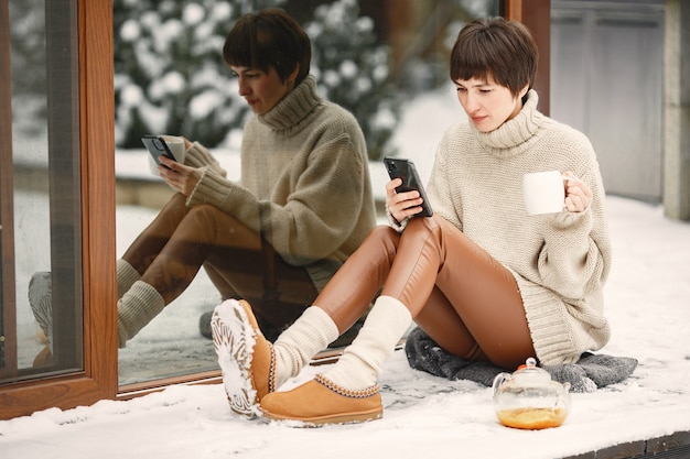 Ritratto del primo piano della donna in maglione bianco, bere il tè e tenere lo smartphone