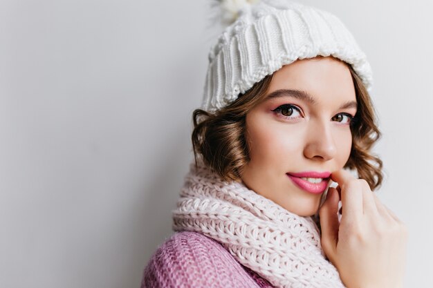 Ritratto del primo piano della donna riccia raffinata in cappello bianco. Ragazza europea estatica con bellissimi occhi in posa in sciarpa carina.