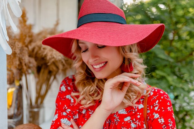 Ritratto del primo piano della donna sorridente bionda alla moda attraente in cappello rosso di paglia e vestito di modo di estate della camicetta con il sorriso sensuale