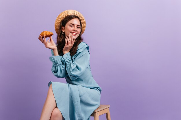 Ritratto del primo piano della ragazza affascinante che posa con il croissant. Modello in boater e vestito di seta blu sorridente carino.