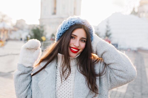 Ritratto del primo piano della ragazza allegra con capelli neri lunghi che posano nella mattina di inverno sulla città della sfuocatura. Signora castana in berretto blu che gode del servizio fotografico in una giornata fredda.