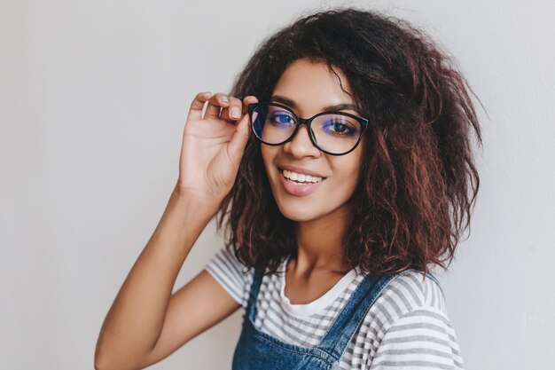 Ritratto del primo piano della ragazza allegra con il sorriso di hollywood e capelli ricci scuri guardando attraverso gli occhiali alla moda