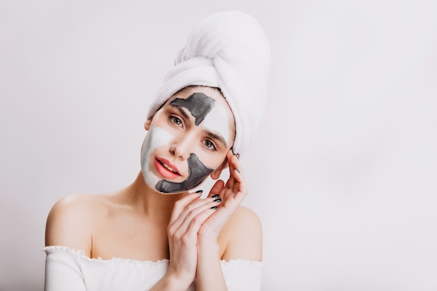 Ritratto del primo piano della ragazza attraente che fa maschera facciale prima di coricarsi. Donna adulta in posa sul muro bianco.