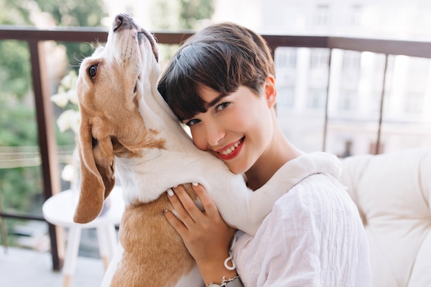 Ritratto del primo piano della ragazza beata con gli occhi grigi in posa con un sorriso felice mentre il suo cane beagle alzando lo sguardo